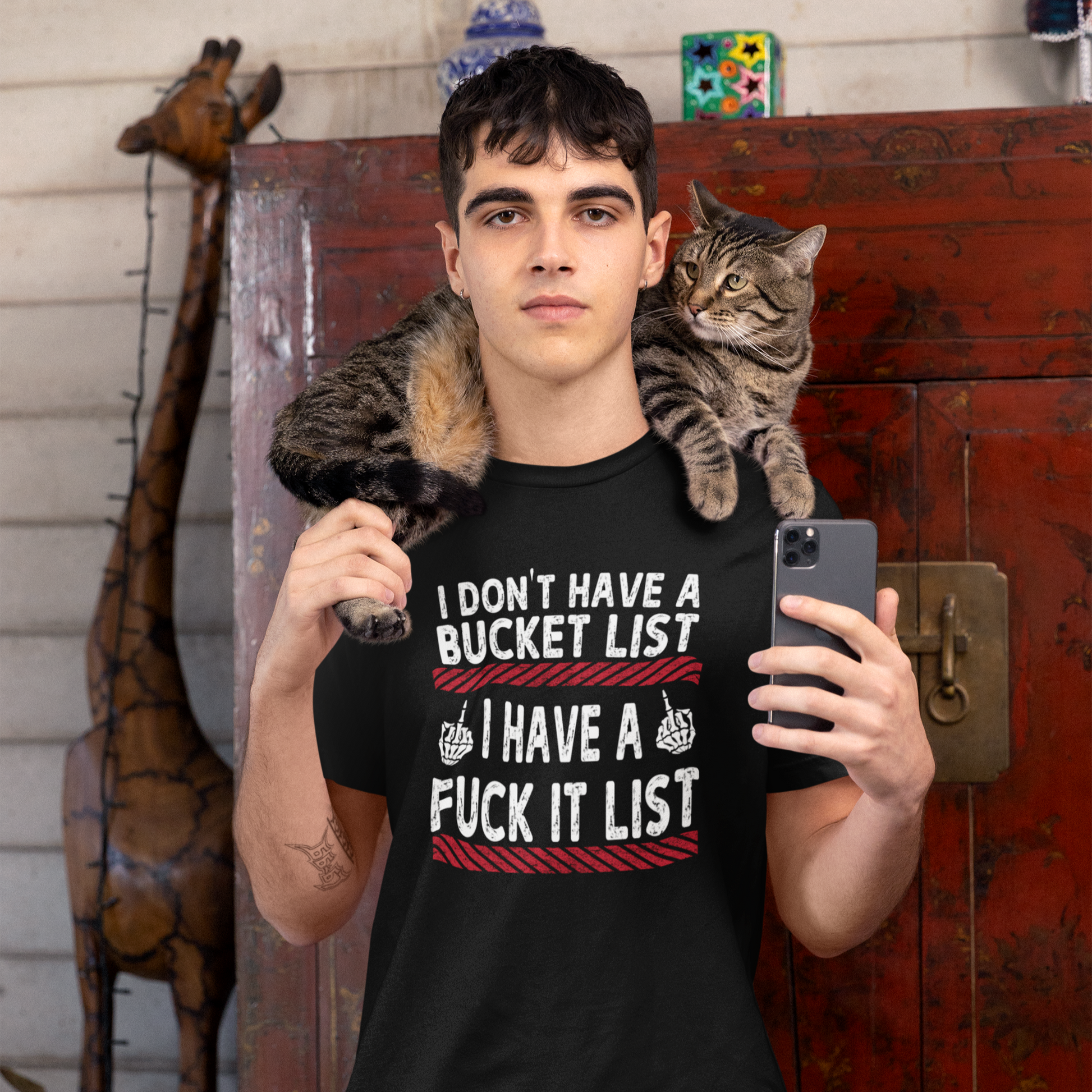 Person wearing a black t-shirt with a humorous design, holding a phone, with a cat on their shoulder.