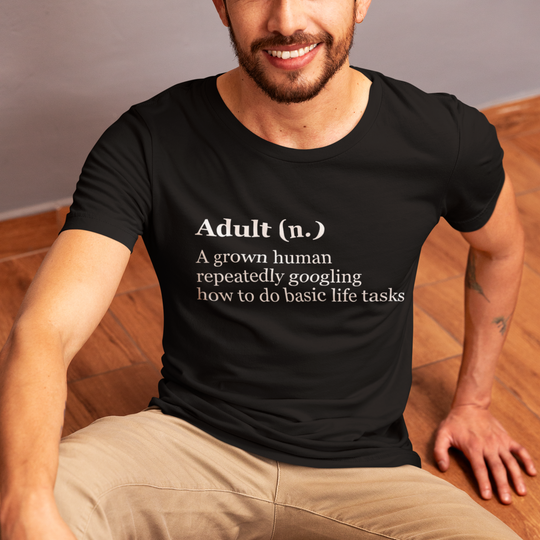 Man wearing a black t-shirt with a humorous definition of 'adult' sitting on a wooden floor.