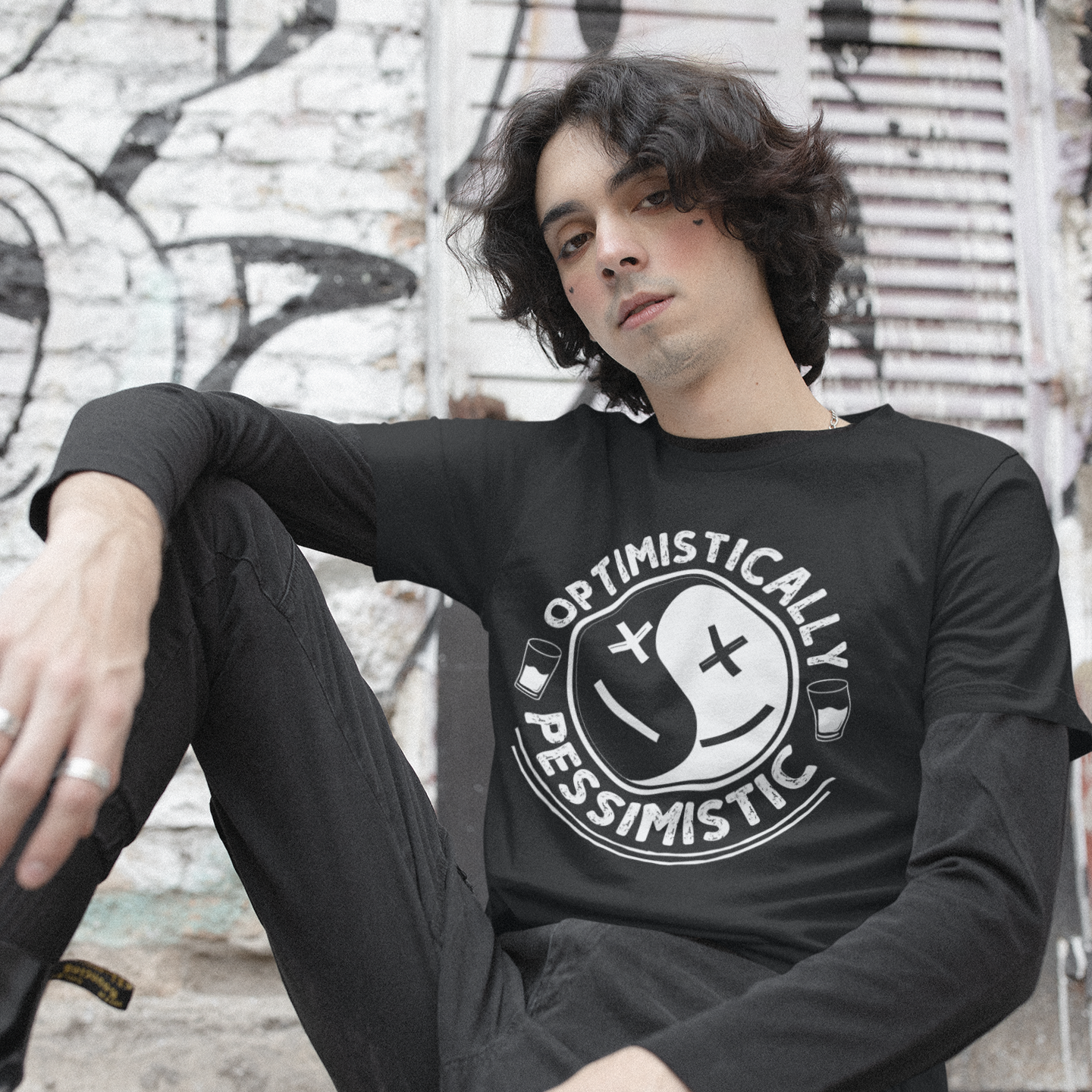 Person wearing a black sweatshirt with 'Optimistically Pessimistic' design, sitting against a graffiti-covered wall.