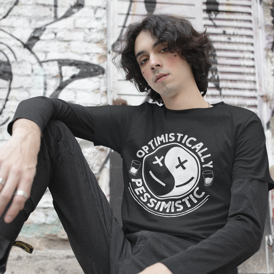 Person wearing a black sweatshirt with 'Optimistically Pessimistic' design, sitting against a graffiti-covered wall.